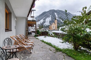 Une terrasse pavée de pierres, avec des chaises et une table en bois, une église dotée d’un clocher, et des montagnes enneigées en arrière-plan.