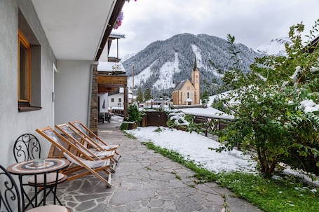 A stone-paved patio with wooden chairs and a table, a church with a steeple, and snow-covered mountains in the background.. Apartment 'Hotel Sonnblick - Wohnung Klaus' with Mountain View, Shared Terrace and Wi-Fi