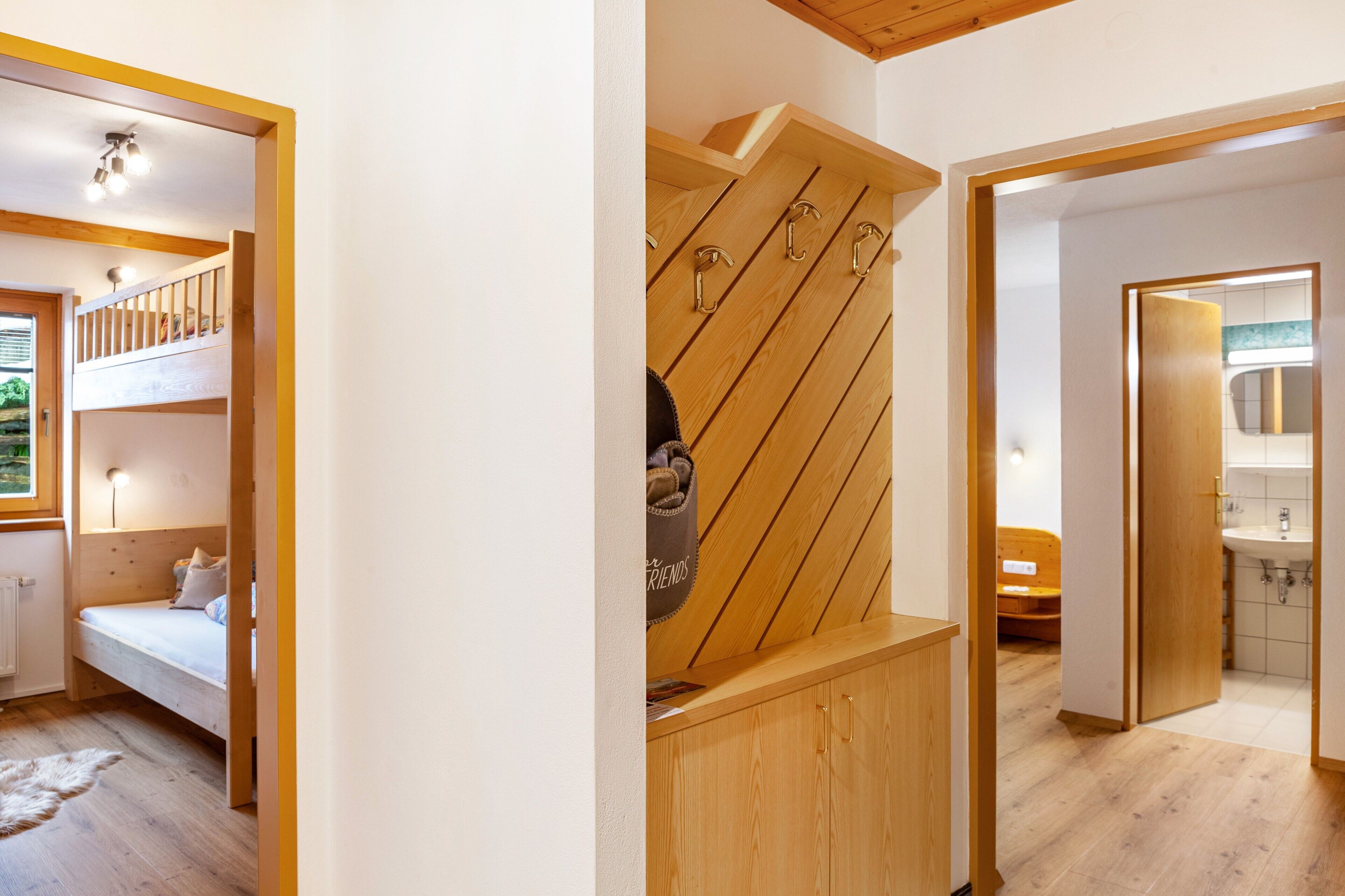 Een kamer met een stapelbed, een houten kast met haken en een badkamer met wastafel en spiegel.