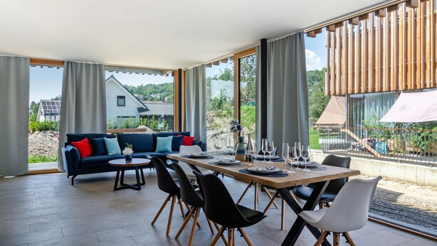 A modern dining area with a table set for four, a sofa with cushions, and large windows offering a view of a house and greenery. - Holiday Home 'Architektenhaus C1 Mit Seesicht' with Lake View, Private Terrace and Wi-Fi (Öhningen)