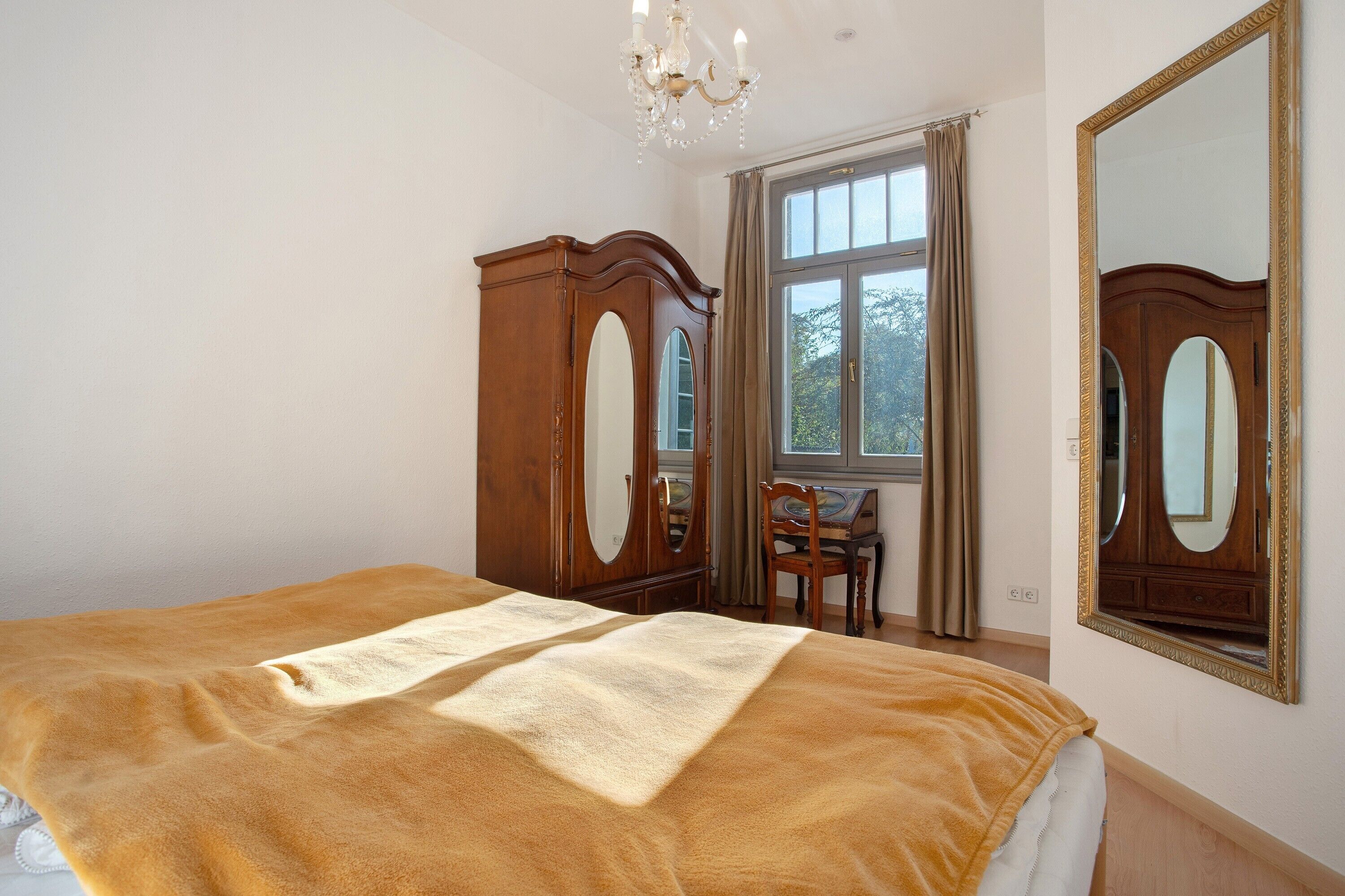 A bedroom with a bed, a wooden wardrobe, a chair, a large mirror, and a window with curtains.