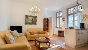 A living room with a sofa, a coffee table, a chandelier, a painting, and a kitchen area with cabinets and a window. - Villa 'Effi-Briest-' with Mountain View, Private Terrace and Wi-Fi (Ostseebad Heringsdorf)