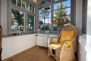 Une véranda avec de grandes fenêtres, un fauteuil en osier avec un coussin jaune et un radiateur.
