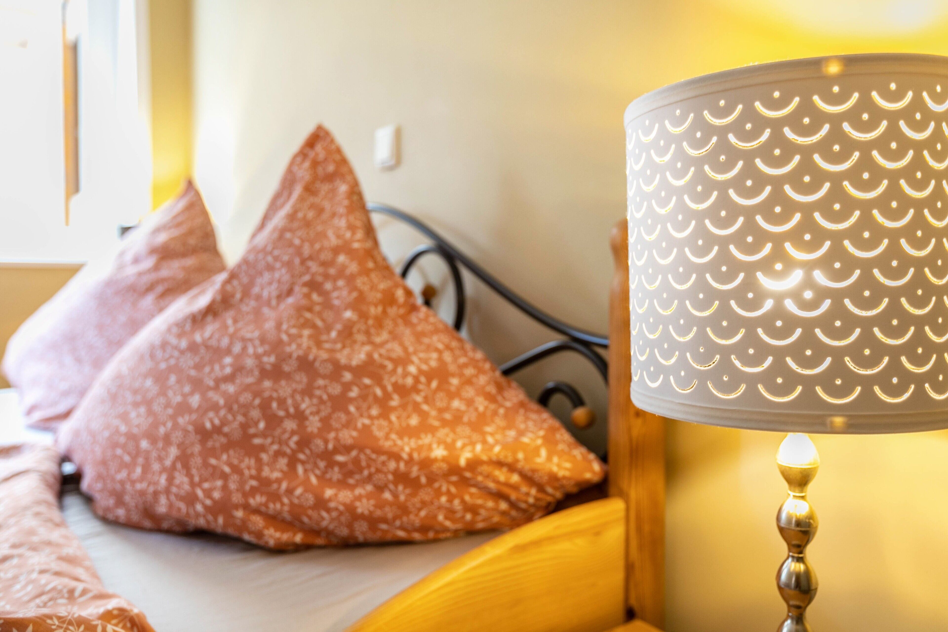 A bed with a patterned pillow, a lamp with a textured lampshade, and a wall socket.