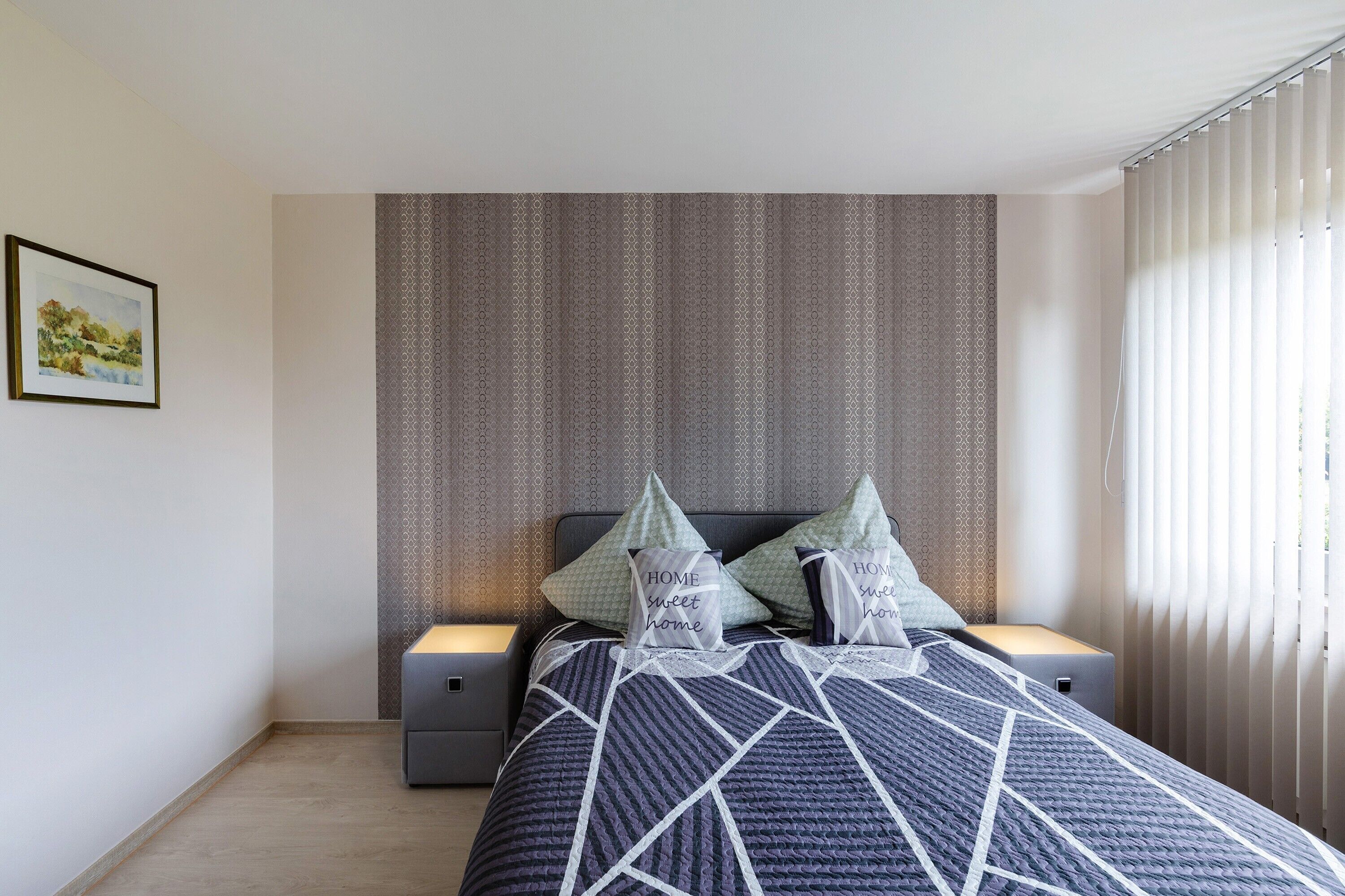 Una cama bien tendida con una colcha de estampado geométrico, dos almohadas con texto, una mesita de noche y un cuadro colgado en la pared.