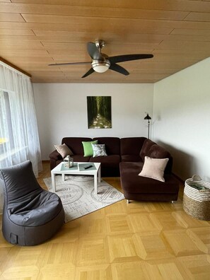 Un salon moderne avec un ventilateur de plafond, un canapé marron, un fauteuil en forme de poire noir, une table basse blanche et un tableau encadré au mur.