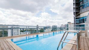 Une piscine sur le toit, dotée d’une terrasse en bois et de rampes en métal, offrant une vue sur un paysage urbain avec des bâtiments modernes et un ciel nuageux.
