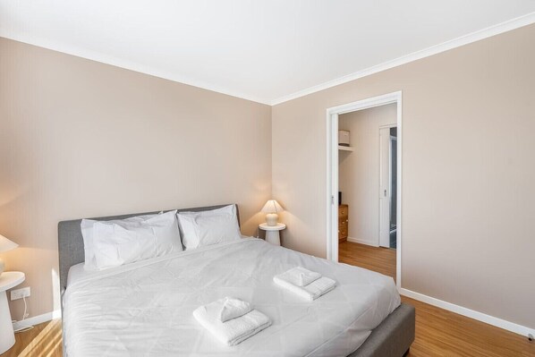 Cama bem arrumada com lençóis e toalhas brancos, uma luminária de cabeceira e piso de madeira.