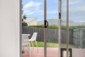 Vista a través de un ventanal hacia un patio trasero con terraza de madera, una silla blanca y una reja metálica.