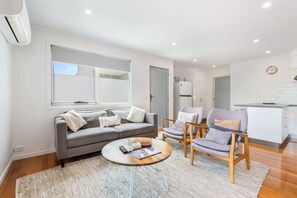Un salon moderne avec un canapé gris, deux fauteuils, une table basse et une cuisine en arrière-plan.