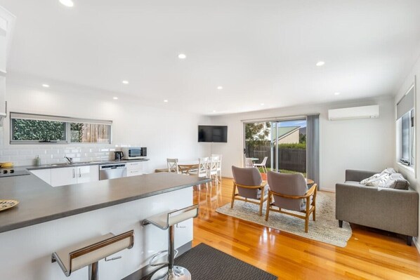 A modern kitchen with a bar area, dining space with chairs, a grey sofa, and a TV mounted on the wall.