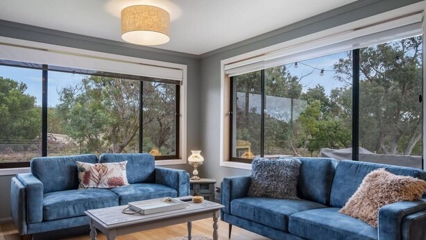 A modern living room with two blue sofas, a coffee table, and large windows offering a view of trees. - Peaceful Beachside Home (Clifton Beach)