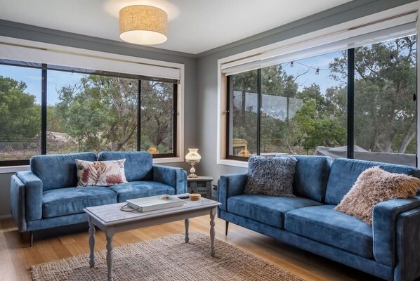 A modern living room with two blue sofas, a coffee table, and large windows offering a view of trees. - Clifton Beach Home on 5.5 Acres, Wood Fire & Deck (Clifton Beach)