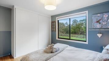 A bedroom with a bed, a window with a view of trees, a wall-mounted light fixture, and a decorative heart hanging on the wall.