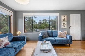 A modern living room with two blue sofas, a coffee table, and a large window with a view of trees. - Peaceful Beachside Home (Clifton Beach)