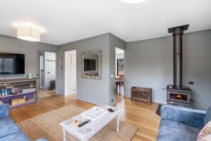 A living room with a fireplace, a television, a bookshelf, and a dining area in the background. - Peaceful Beachside Home (Clifton Beach)