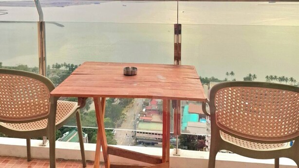 A balcony with a wooden table and two wicker chairs, overlooking a body of water and a distant landscape.