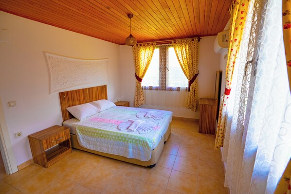 A bedroom with a wooden ceiling, a bed with a patterned bedspread, two windows with curtains, and a nightstand. - Defne Olive Gardens Hotel Fethiye (Fethiye)