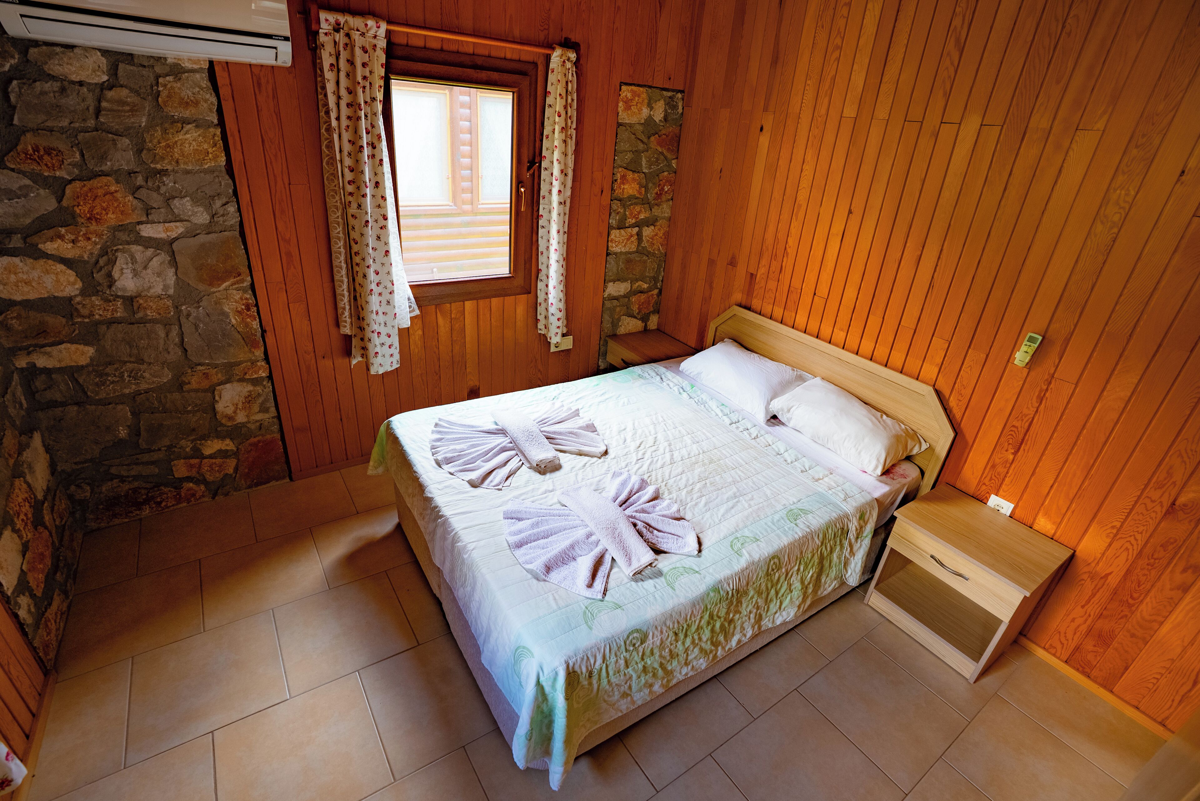 A bedroom with a wooden wall, a stone wall, a window with curtains, a bed with towels, and a nightstand.