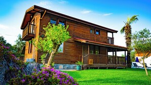 Une maison en bois à deux étages, dotée d’un balcon, entourée d’une pelouse bien entretenue et de plantes en fleurs.