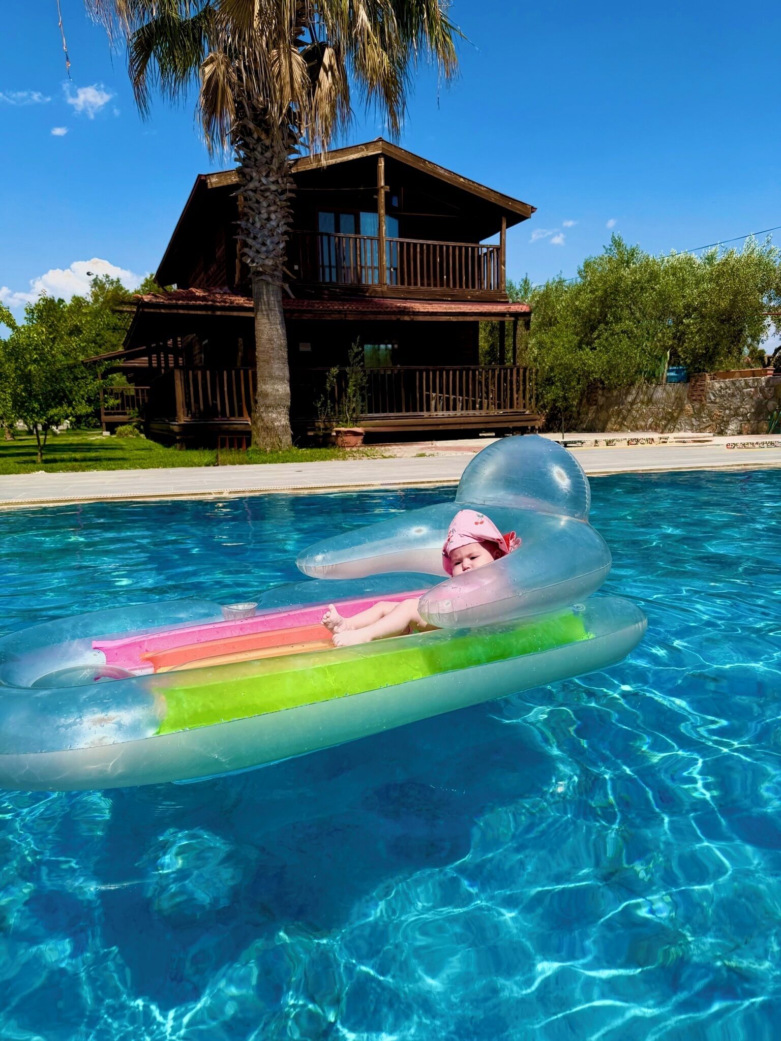 Une piscine avec une eau d’un bleu limpide, une personne flottant sur une bouée gonflable et, au fond, une maison en bois dotée d’un balcon.