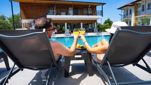 Deux personnes sont assises sur des chaises longues au bord d’une piscine, avec en arrière-plan un bâtiment en bois.