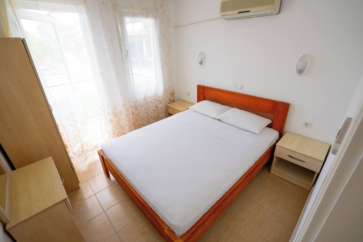 Een slaapkamer met een houten bed, twee nachtkastjes, een raam met gordijnen en een wandairconditioner.