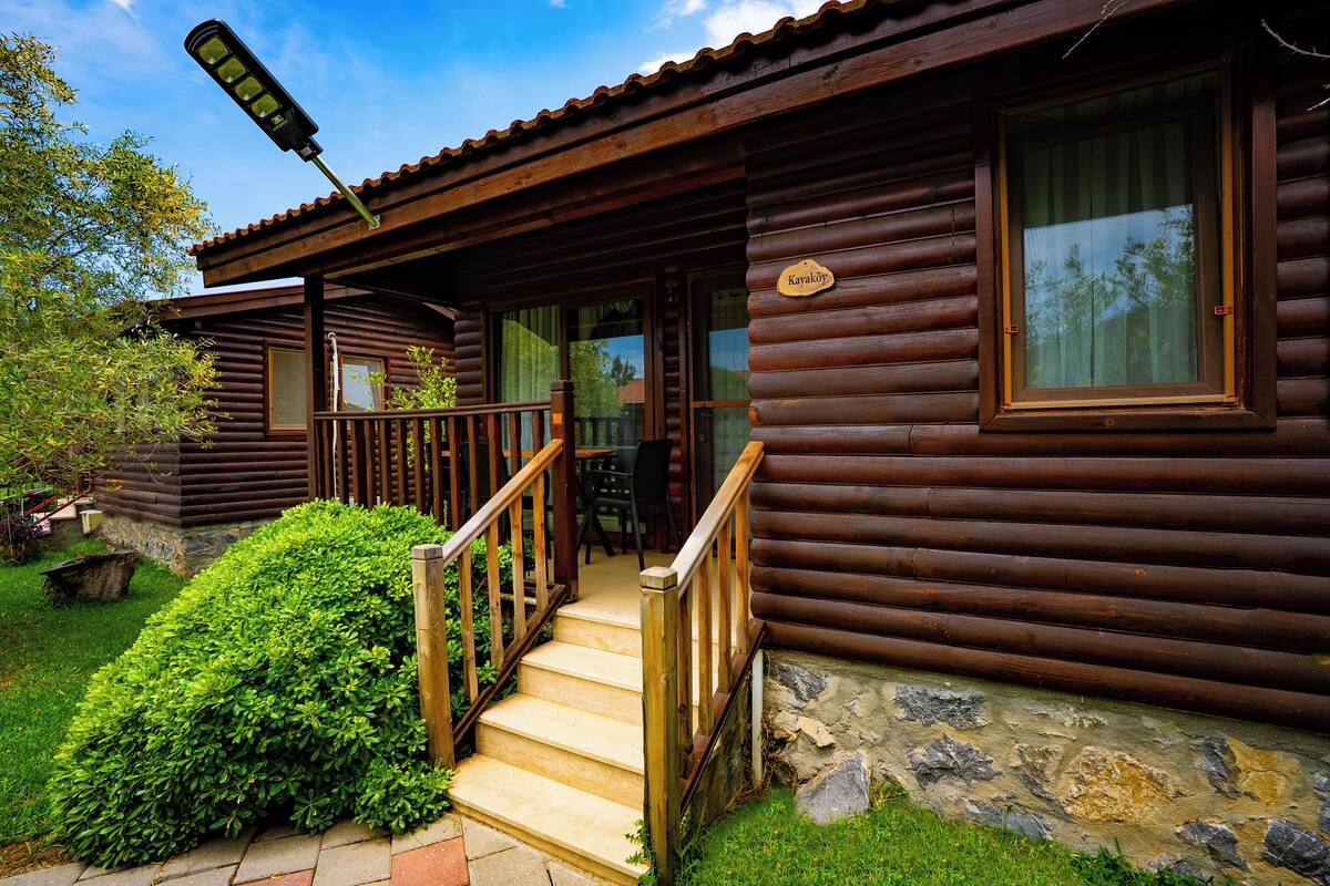 Een vakantiehuis van hout met een houten trap naar een veranda, een bord met de tekst 'Kavakoy' en een stenen fundering.