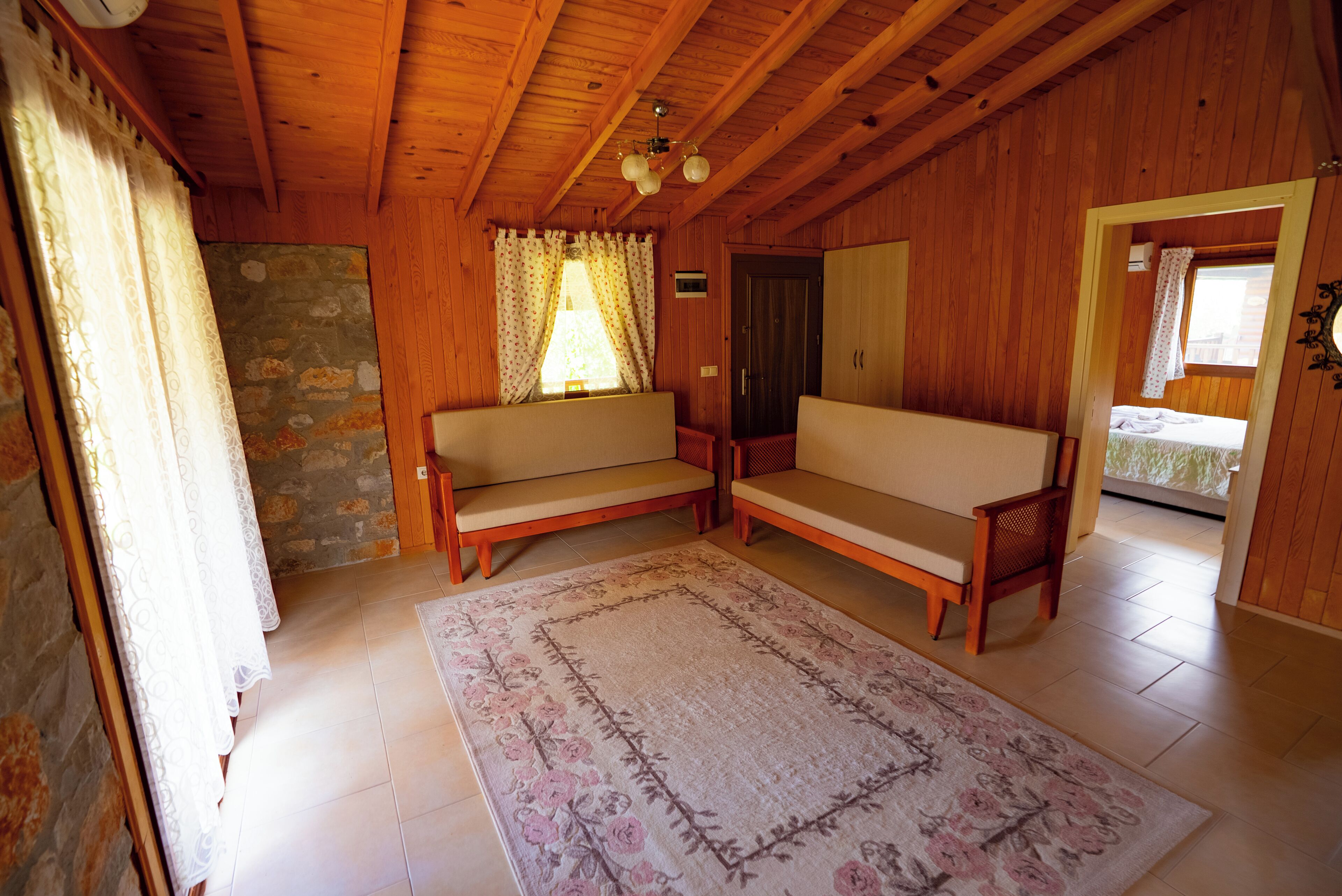 A wooden cabin interior with a stone wall, two wooden sofas, a floral rug, and a ceiling fan.
