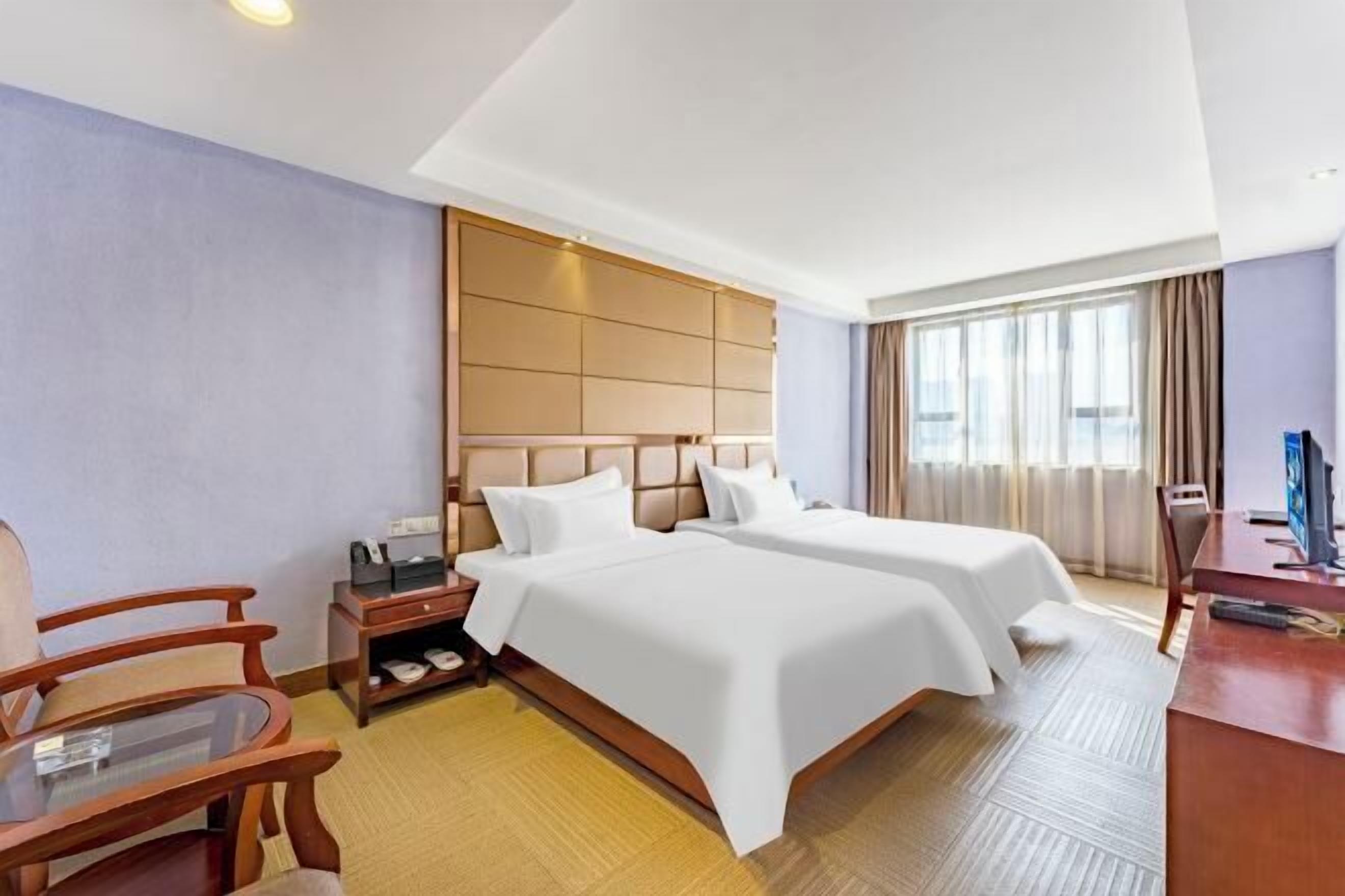 A hotel room with two beds, a desk, a chair, a nightstand, and a window with curtains.