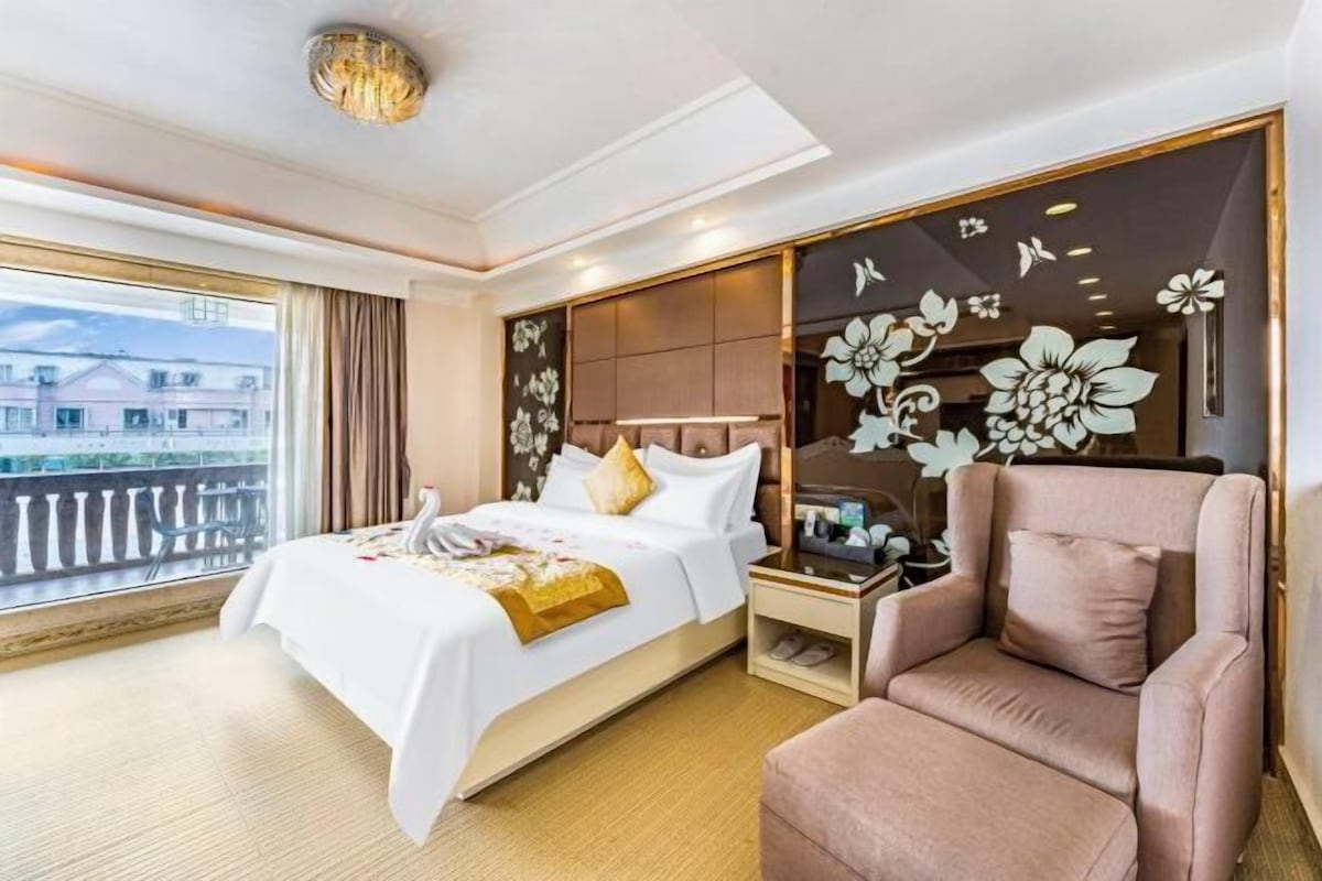 A hotel room with a bed, a chair, a nightstand, a decorative wall, and a view through a window.