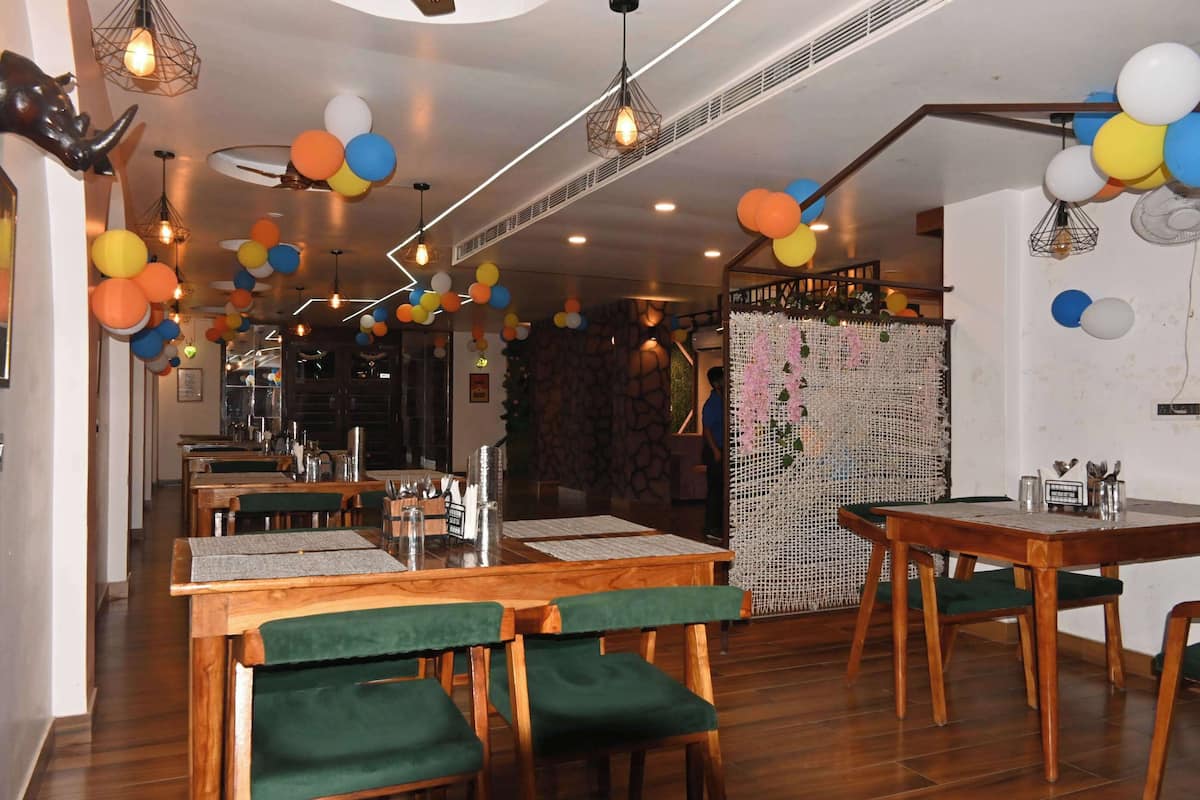 Interior de restaurante con mesas de madera, sillas con cojines verdes, pared de piedra y techo decorativo con lámparas colgantes.