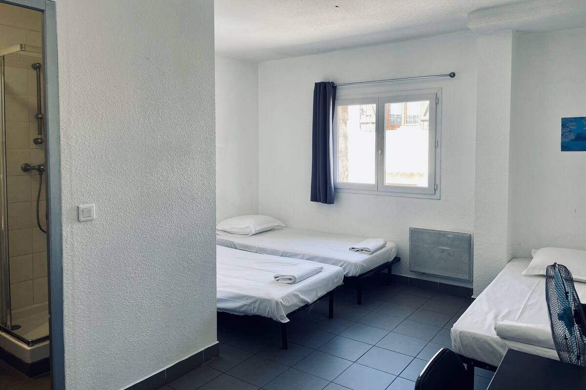 Une petite pièce propre, avec deux lits, une fenêtre avec des rideaux et un espace douche.