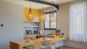 A modern kitchen with wooden cabinetry, a central island with seating, pendant lighting, and a window with sheer curtains. - Ambhara X Apartment by Betterplace (Kerobokan)