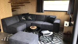 Ein modernes Wohnzimmer mit einem grauen Ecksofa, zwei schwarzen Couchtischen, einem gemusterten Teppich und einem Fenster mit Vorhängen.