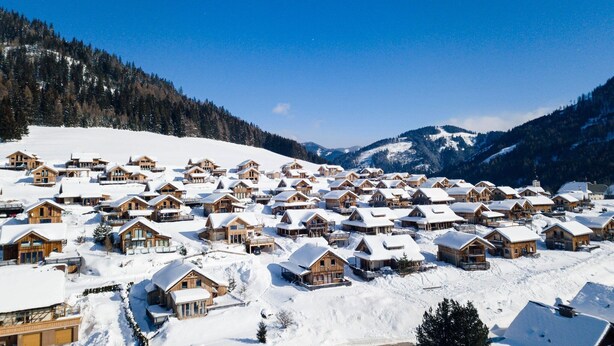 Ein schneebedecktes Dorf mit Holzhütten, umgeben von Bergen und blauem Himmel.