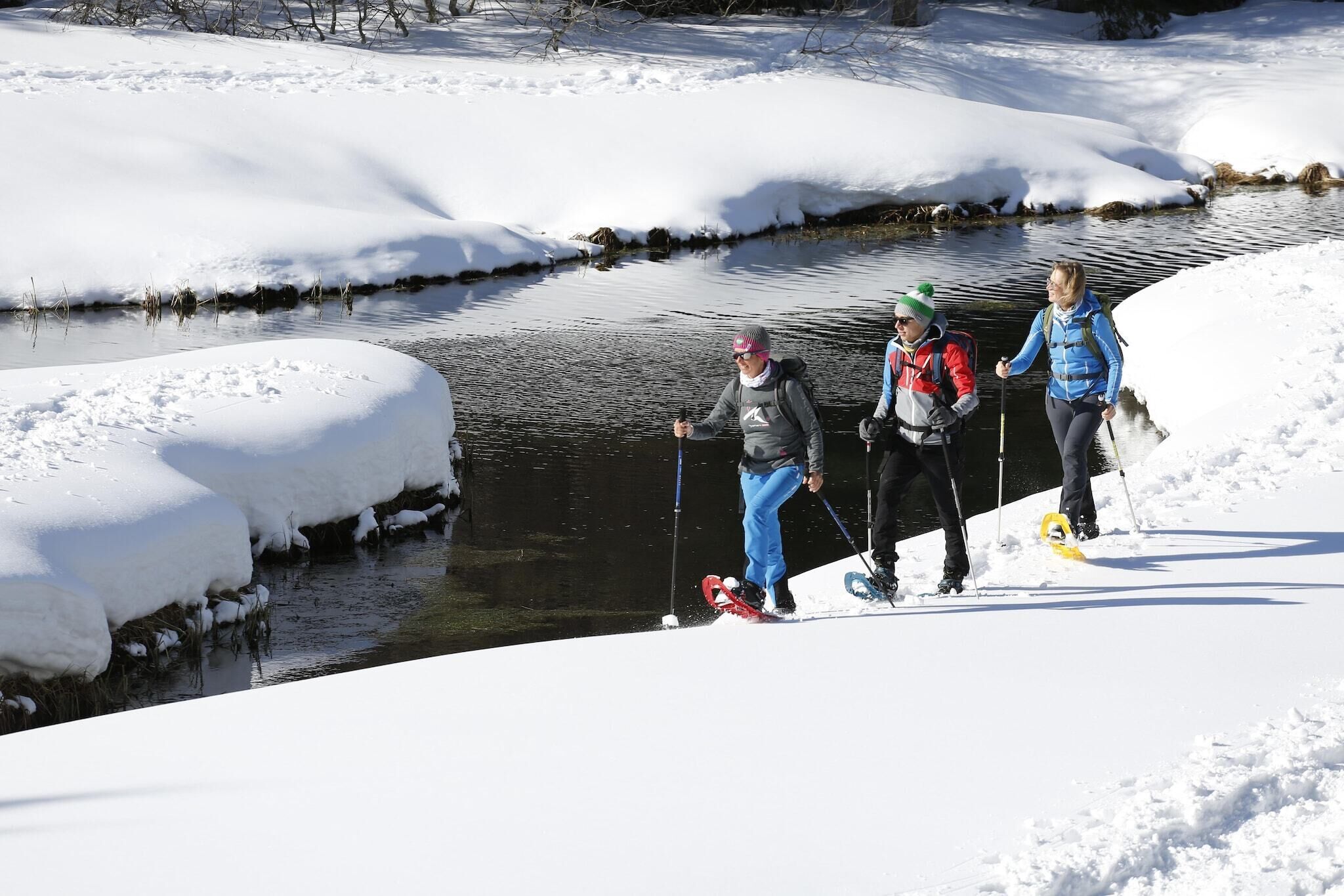 A snowy landscape with a river, people skiing, and a bridge.