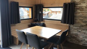 Ein Eckzimmer mit Holztisch und -stühlen, großen Fenstern mit Vorhängen und einer Steinwand.