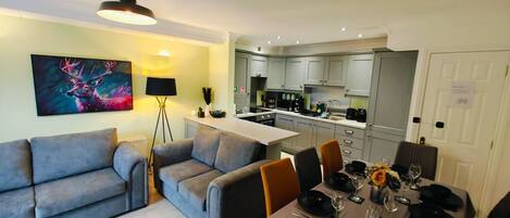 Uma sala de estar moderna com um conjunto de sofá cinza, uma mesa de jantar posta para quatro pessoas e uma cozinha ao fundo.