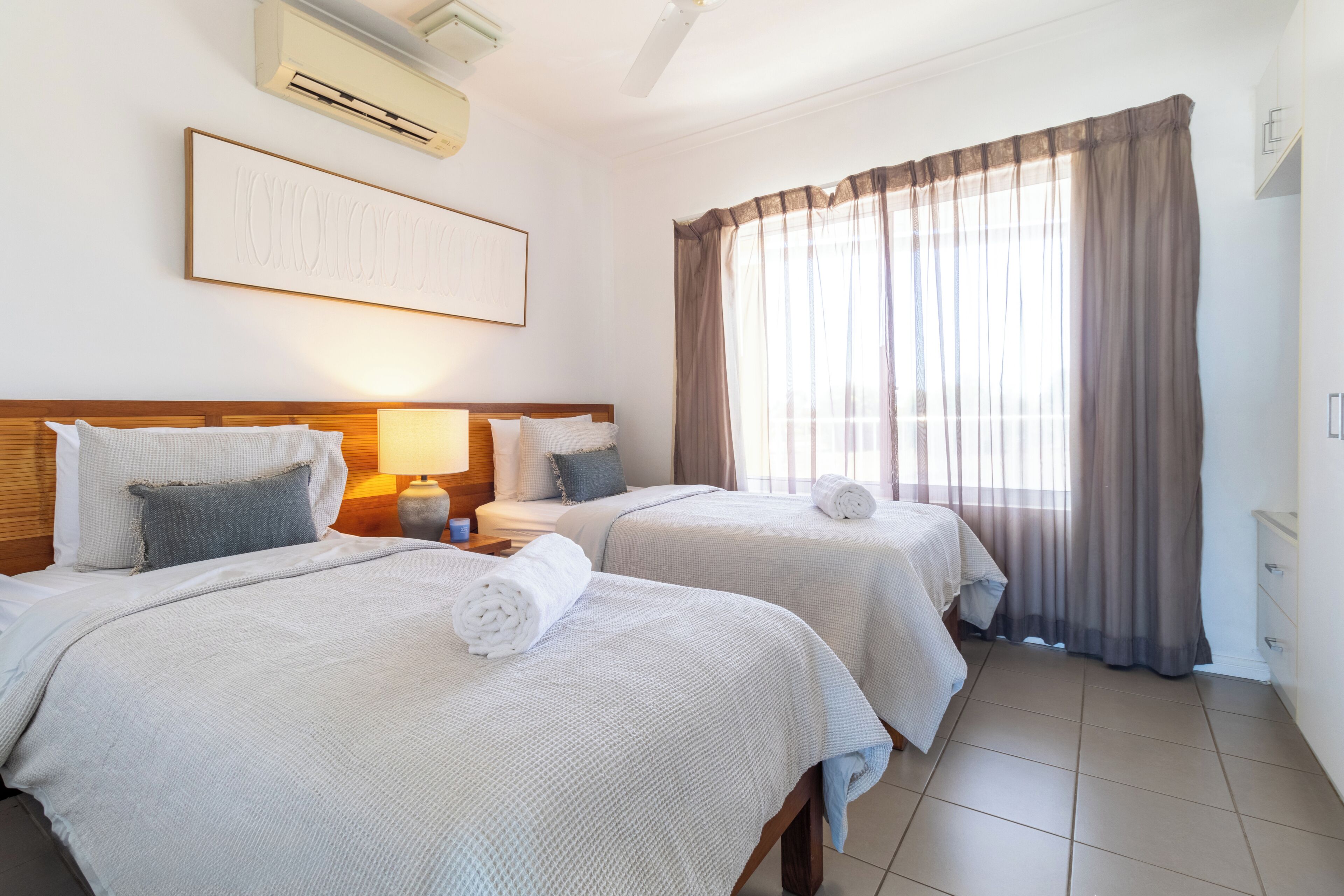 A hotel room with two beds, a large window with curtains, a wall-mounted air conditioner, and a framed picture.