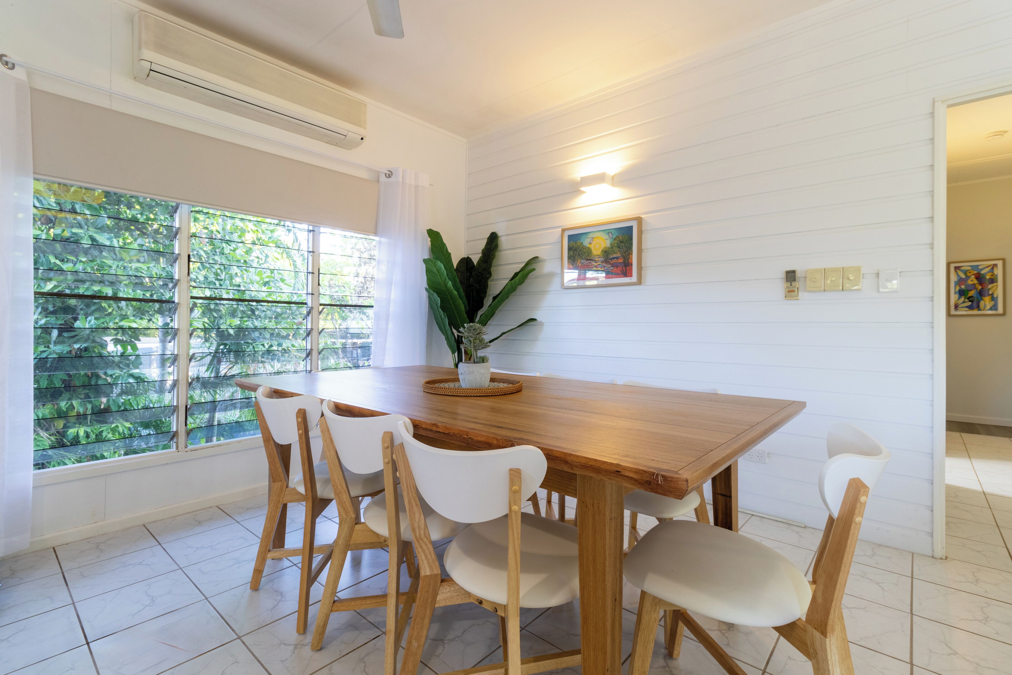 Uma zona de refeições com uma mesa de madeira, cadeiras brancas, um vaso de planta e uma janela com vista para vegetação.
