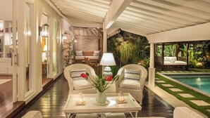 A cozy indoor seating area with wicker furniture, a table with a vase and candles, and a view of a pool outside. - Villa Karina Umalas (Kerobokan)