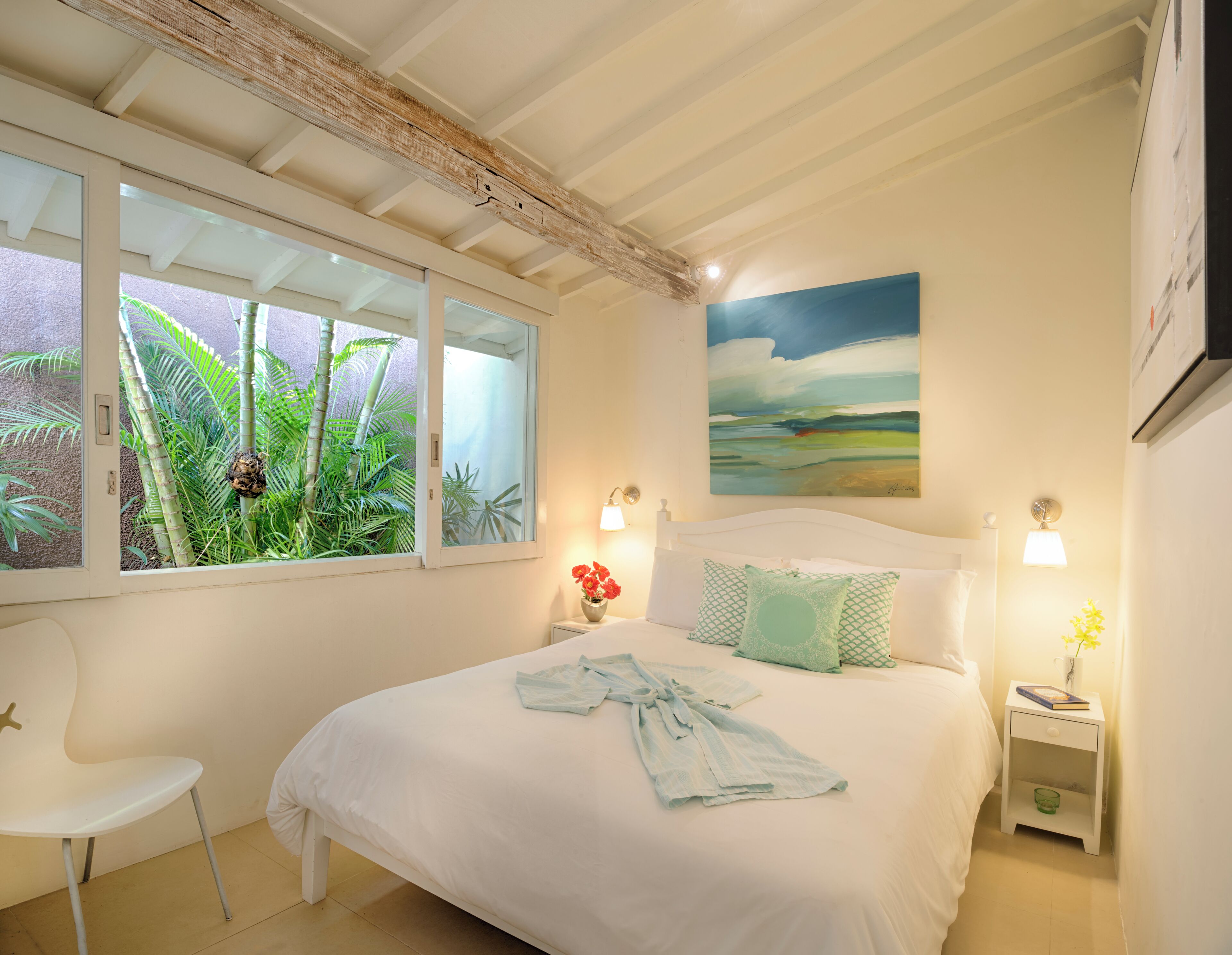 A bedroom with a bed, a nightstand, a chair, a window with a view of greenery, and a painting on the wall.