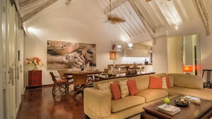A spacious living room with a sofa, dining table, and chairs. There is a kitchen in the background with cabinets and a clock. - Villa Karina Umalas (Kerobokan)