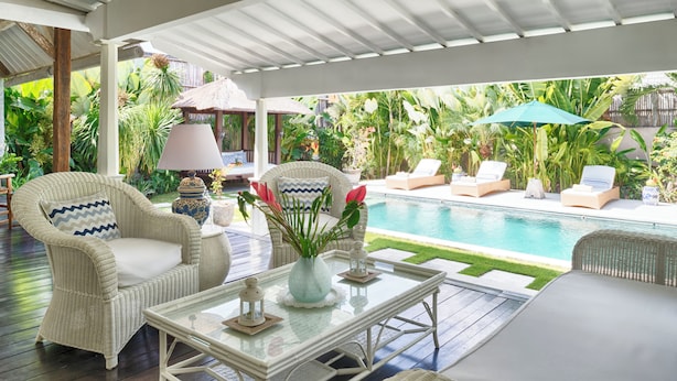 A covered outdoor seating area with wicker furniture, a glass table, and a pool in the background. - Villa Karina Umalas (Kerobokan)
