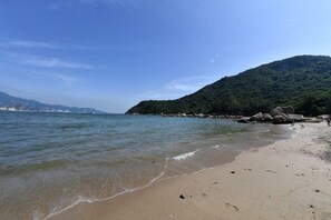Ein Strand mit klarem Wasser, einem sandigen Ufer und einer bewaldeten Hügelkette im Hintergrund.