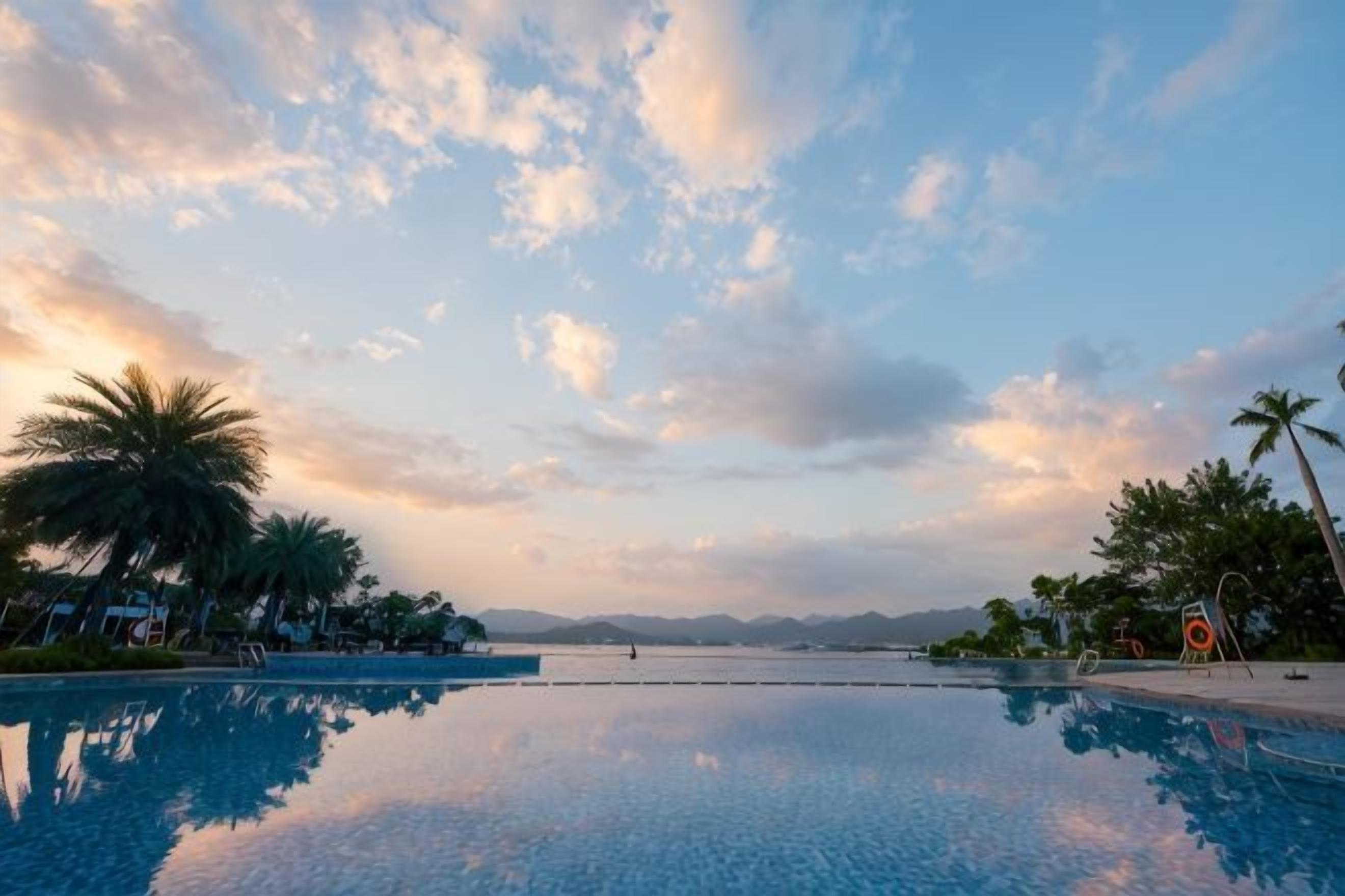 Cielo despejado con nubes, una piscina tranquila que refleja el entorno, y palmeras a la izquierda.