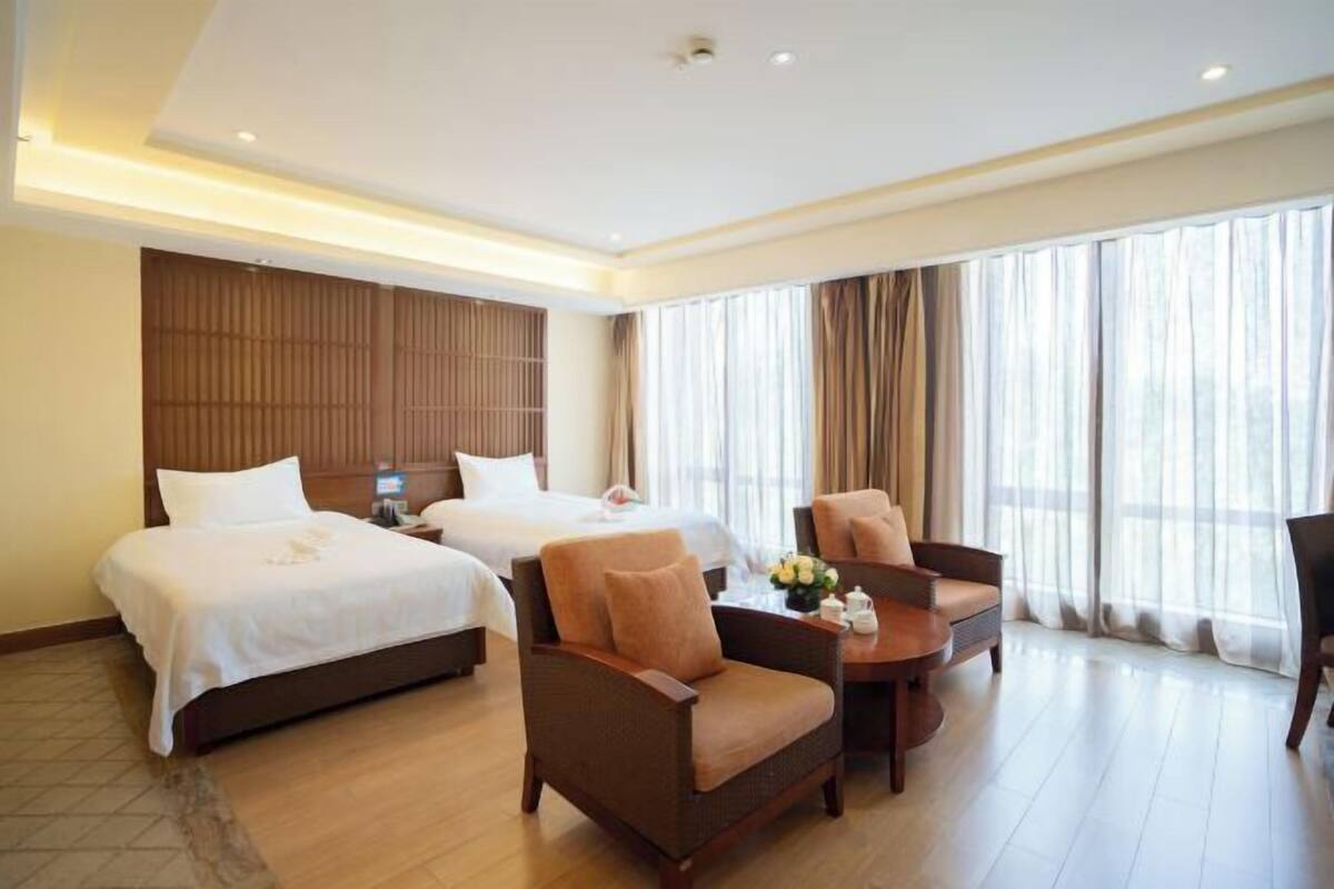Una habitación de hotel con dos camas, un área de estar con una mesa y sillas, y ventanas amplias con cortinas.