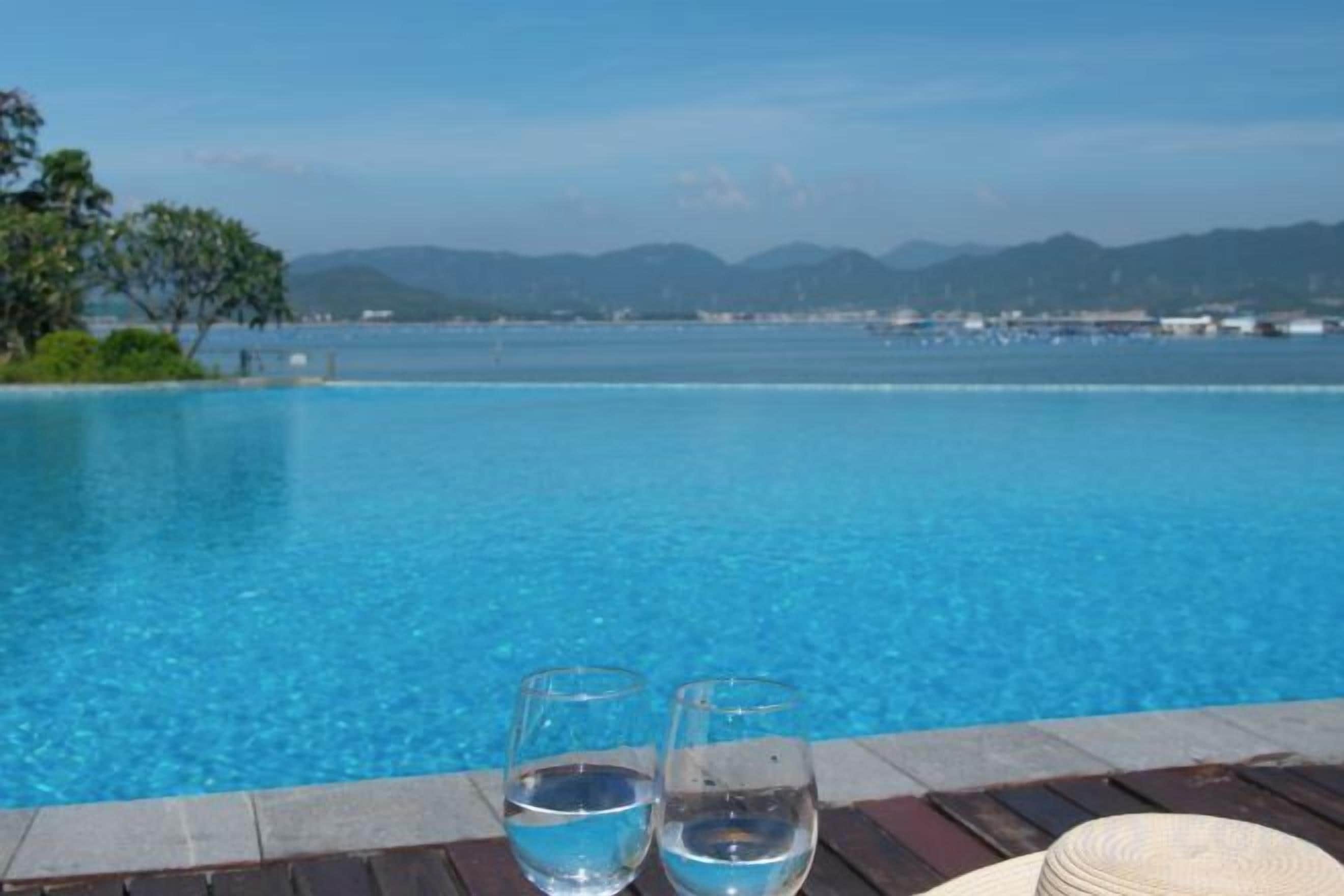 A clear blue pool with two glasses of water on a wooden deck, overlooking a body of water and distant mountains.