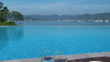 A clear blue pool with two glasses of water on a wooden deck, overlooking a body of water and distant mountains.